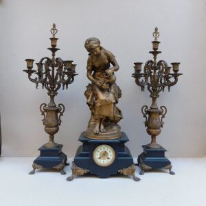Ρολόι μεταλλικό με άγαλμα κοπέλας, μαρμάρινη βάση και δυο πεντάκερα κηροπήγια. Είναι γαλλικό κατασκευασμένο το 1855.